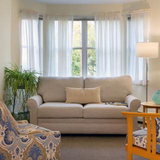 A cozy living room with a beige sofa, patterned armchair, and a floor lamp by the window.