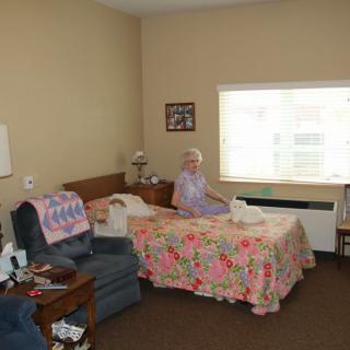An elderly person sitting on a bed in a nursing home room with cozy furnishings.