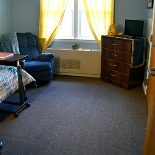 A cozy dorm room with a bed, chair, and dresser near a window with yellow curtains.