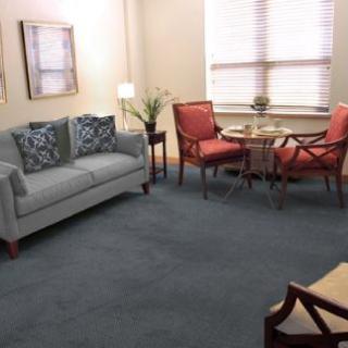 A cozy living room with a sofa and chairs around a small table, featuring soft lighting and a carpeted floor.