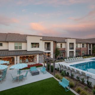 A modern residential complex featuring a large swimming pool and outdoor seating area under a beautiful sunset sky.