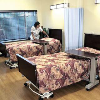 A caregiver tidies up a shared room in a nursing home, with neatly arranged beds and bedside tables.