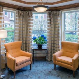 A cozy interior space featuring two orange armchairs flanking a small table with a potted plant, set against large windows with curtains.