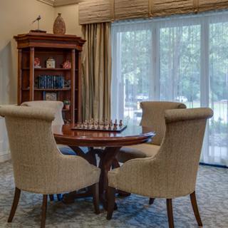 A cozy dining room with a chess set on the table, surrounded by upholstered chairs and a bookshelf in the corner.