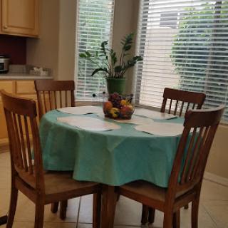 A cozy dining area featuring a round table with a turquoise tablecloth and a centerpiece of fruits, complemented by the warmth of natural light.
