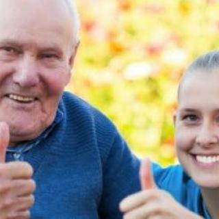 An elderly man and a young woman smiling and giving a thumbs up outdoors.