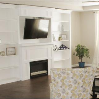 A cozy living room with a television mounted above a fireplace, surrounded by white shelves and decor.
