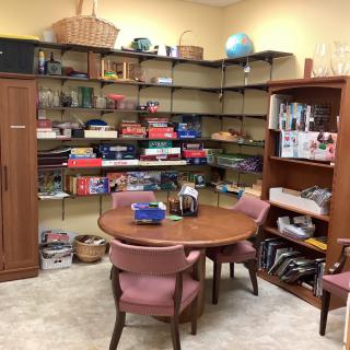 A cozy office space with a round table, chairs, and well-organized shelves filled with books and board games.
