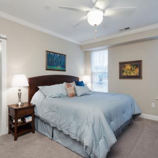 A cozy and well-lit bedroom featuring a large bed with blue bedding, flanked by nightstands with lamps, a ceiling fan, and paintings on the walls.