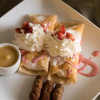 A delicious plate with strawberry crepes topped with whipped cream and served alongside breakfast sausages.
