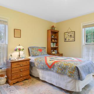 A cozy bedroom with a single bed featuring a colorful quilt, wooden nightstand and bookshelf, illuminated by natural light from two windows.