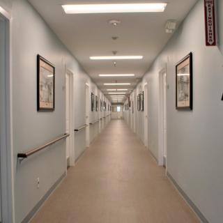 A long, well-lit corridor in a hospital or clinic with doors and framed pictures on the walls.