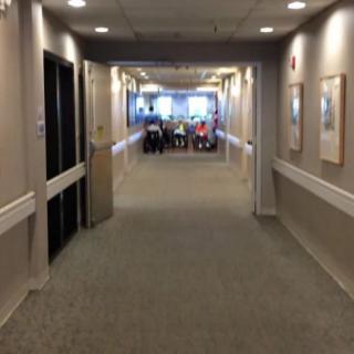 A carpeted hallway with overhead lighting, leading to a room with chairs and people in the distance.