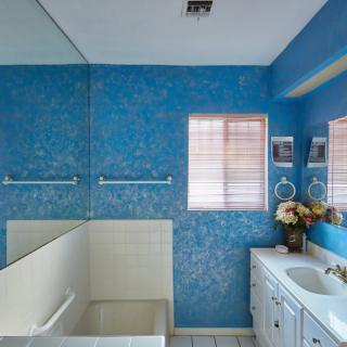 A brightly lit bathroom with blue patterned walls, a large mirror, and a white sink cabinet.