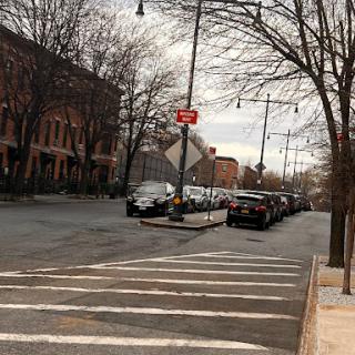A quiet urban street scene featuring parked cars, a crosswalk, and traffic signs against an overcast sky.
