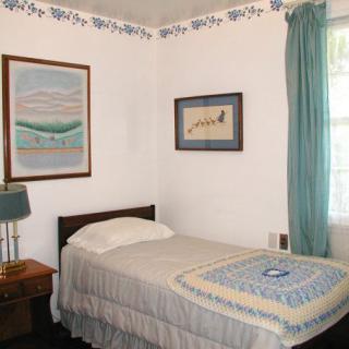 A small bedroom with a single bed, decorated with paintings and a quilt, featuring a window with curtains.