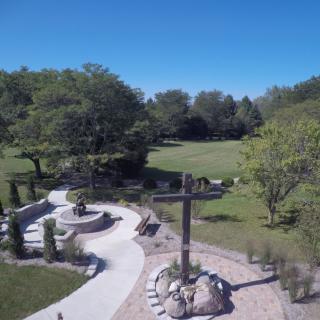 A serene outdoor pathway featuring a large cross, surrounded by trees and greenery.