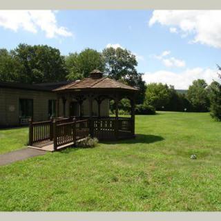 A scenic garden with a wooden gazebo surrounded by a lush green lawn and trees under a partly cloudy sky.