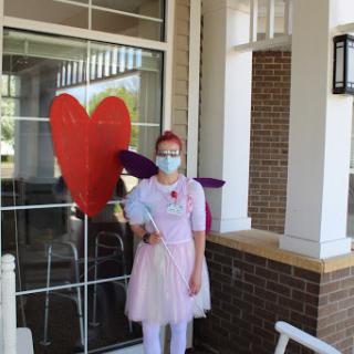 A person in a fairy costume holding a heart-shaped decoration, spreading joy outdoors while wearing a mask.