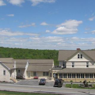 A scenic view of a large house with vehicles parked in front, set against a backdrop of trees and a partly cloudy sky.