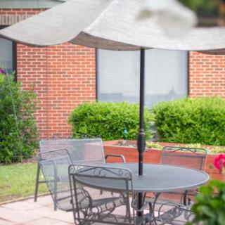 A cozy outdoor patio with a round table and umbrella, surrounded by greenery and a brick building in the background.