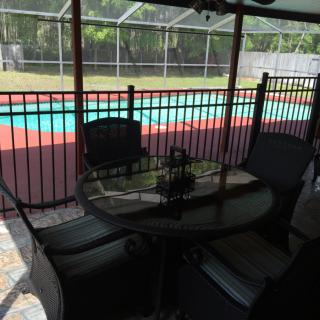 A peaceful backyard scene featuring a swimming pool, bordered by a fence, with patio furniture set under a covered area.