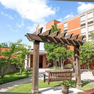 A serene garden area with a wooden pergola and bench, set against a backdrop of trees and a brick building under a clear blue sky.