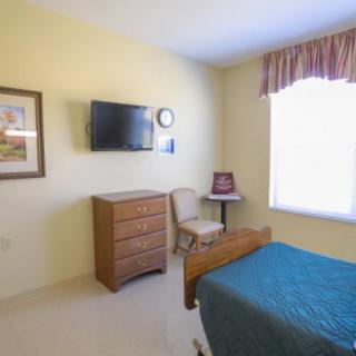 A small bedroom with a television, dresser, chair, and a hospital bed next to a window.