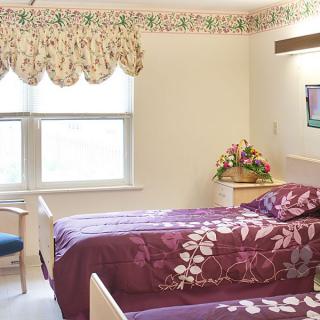 A cozy bedroom with two beds featuring purple floral bedding, a window with decorative curtains, and a vase of flowers on a nightstand.