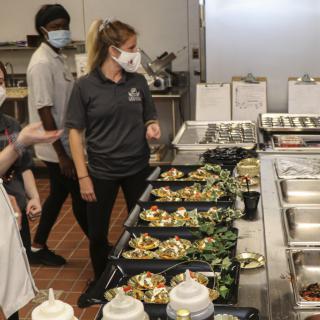 People working in a commercial kitchen preparing meal trays while wearing masks.