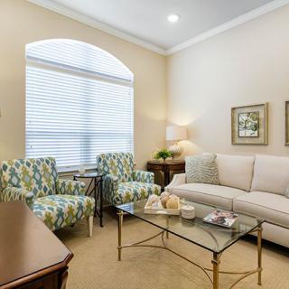A cozy living room featuring a sofa, patterned armchairs, and a glass coffee table with decorative items. A large window allows natural light to illuminate the space.