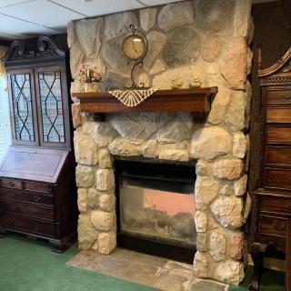 A rustic stone fireplace with a wooden mantel, flanked by vintage wooden furniture in a cozy living room setting.