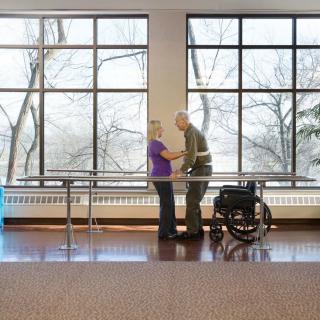 An elderly person receiving physical therapy assistance in a spacious room with large windows.