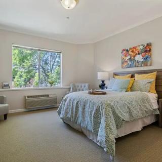 A cozy bedroom with soft lighting, featuring a neatly made bed, artwork on the wall, and a large window letting in natural light.