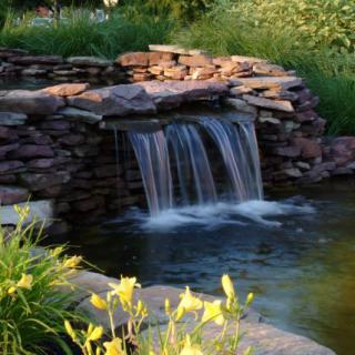 A tranquil garden pond with a small waterfall cascading over a stone wall, surrounded by lush greenery and blooming flowers.