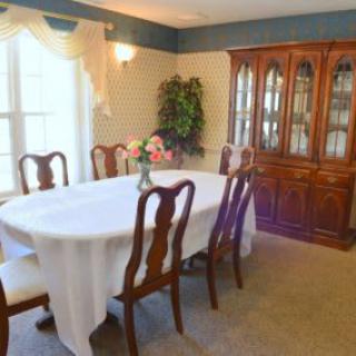 A traditional dining room with a table covered by a white cloth, surrounded by wooden chairs and featuring a glass-fronted cabinet.