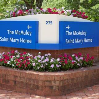 A blue sign for The McAuley Saint Mary Home surrounded by flowers and a brick wall.