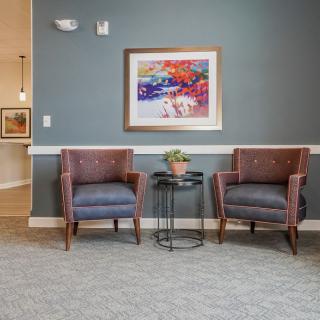 A cozy waiting area featuring two upholstered chairs, a small table with a plant, and colorful artwork on the wall.