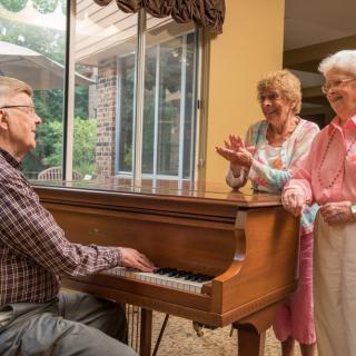 A group of elderly friends enjoying music, with one person playing the piano while others smile and clap indoors.