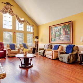 A spacious living room with warm yellow walls, comfortable armchairs, and a large wall painting under abundant natural light from tall windows.