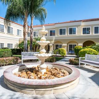 A sunny courtyard featuring a central fountain, surrounded by benches and greenery, with a building in the background.