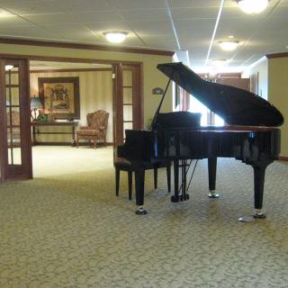 A grand piano is situated in a carpeted living room with an open doorway leading to a cozy seating area.