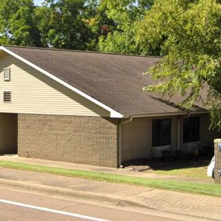 A suburban residential house with a sloped roof, surrounded by trees and located next to a street.