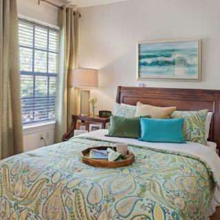 A cozy and well-decorated bedroom featuring a bed with patterned bedding, colorful pillows, a bedside table with a lamp, and a window with curtains. A painting hangs above the bed.