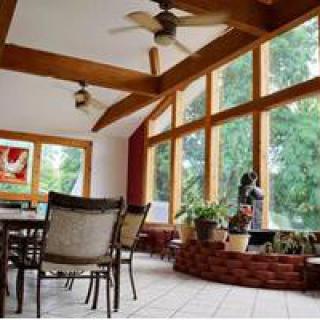 A bright sunroom featuring a dining area with large windows, wooden beams, and ceiling fans, creating a cozy space filled with natural light and greenery.