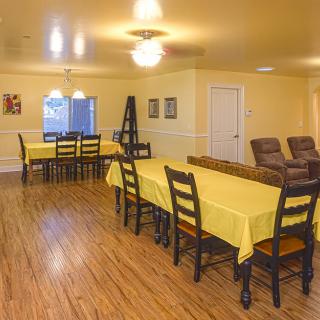 A dining room with tables covered in yellow tablecloths and wooden chairs on a wood floor.