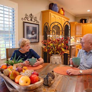 A couple enjoying a conversation over coffee at their kitchen table, surrounded by vibrant home decor and a bowl of fresh fruit.