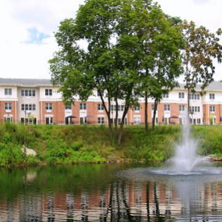 A serene view of a residential building beside a pond featuring a fountain and lush greenery.