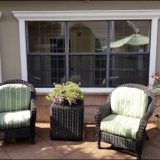 Two wicker chairs with green cushions on a patio, flanking a small plant stand in front of a large window.