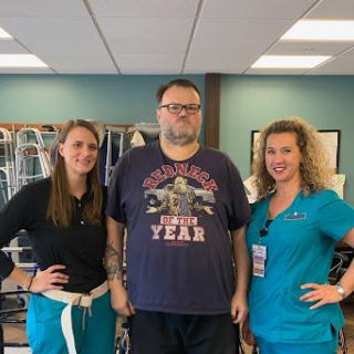 A patient stands proudly with two healthcare professionals during a rehabilitation session.